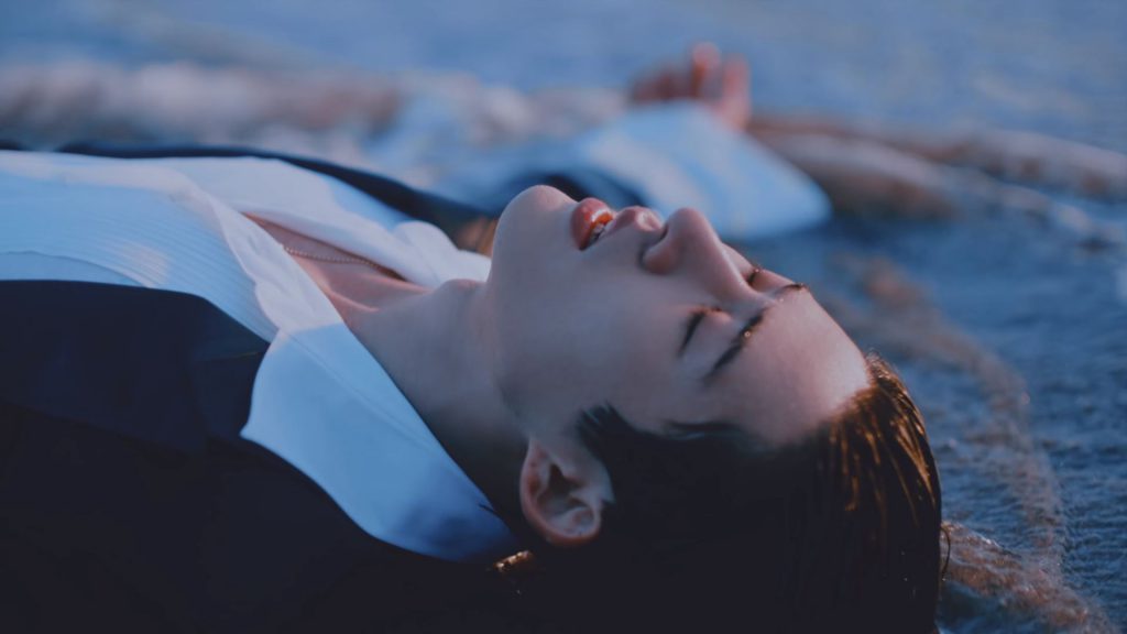 Solo singer and artist B.I laying on the beach as waves crash against him in his "illa illa" music video under 131LABEL.
