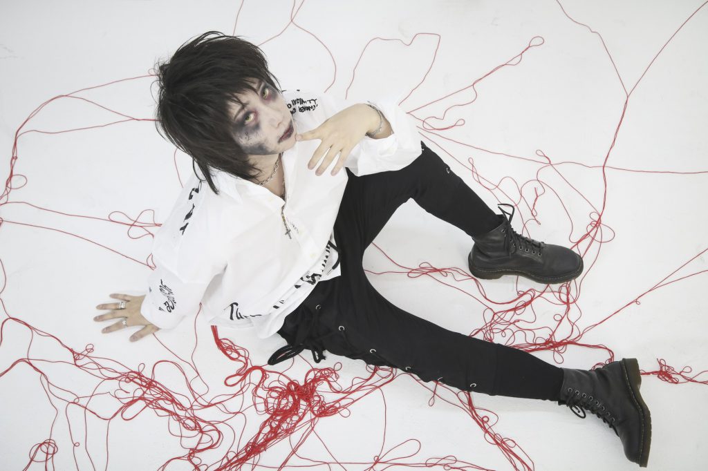 Photo of VOID's bassist Suhyun wearing a white and black outfit sitting on a white floor surrounded by red string.